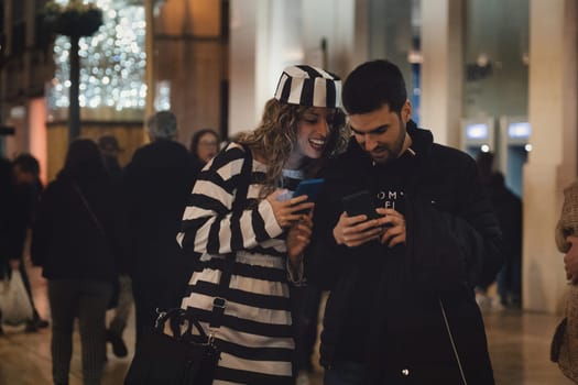 woman and a man standing close together looking at a smartphone, which the man is holding. The woman is smiling. She is wearing a striped garment that is black and white, and also a hat with similar stripes. The man is wearing a dark jacket. High quality photo