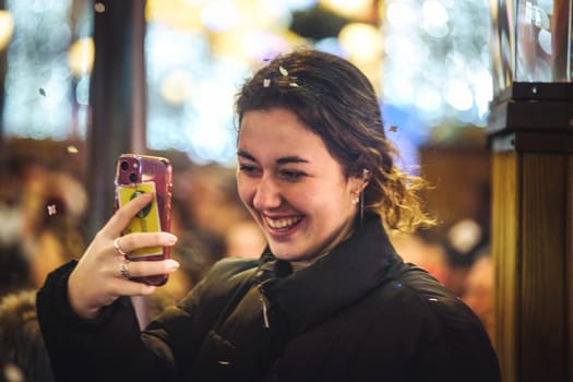 smiling young woman holding up a smartphone, taking a photo or a selfie. She appears to be in a well-lit, indoor environment with a bokeh effect in the background created by various light sources. festive setting, She's dressed in a black coat