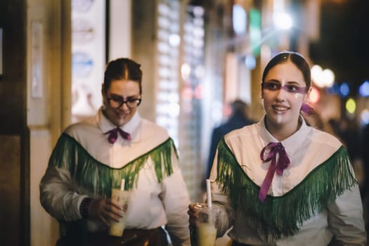 Women dressed in traditional or costume attire walking. both wearing white shirts with green fringe details on the shoulders and purple neckties. smiling and looking towards the camera. Each of them is holding a beverage
