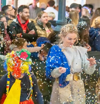 group of people during what appears to be a festive occasion or celebration, as evidenced by the confetti in the air and the cheerful expressions on their faces. In the center is a woman wearing a crown-like accessory on her head and a smile on her face, which suggests she might be part of a special event or ceremony
