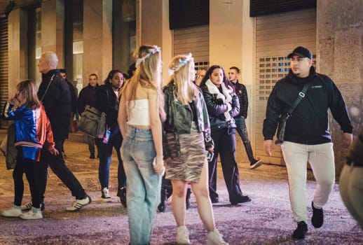 city street at night. Some people are standing and appear to be talking amongst themselves, while others are walking, likely passing by. The environment an urban setting with buildings lining the street