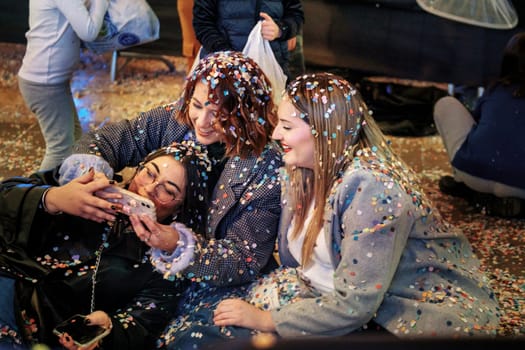 three woman taking a selfie together. They appear to be in a celebratory or festive environment, as they are covered with colorful confetti. The mood joyful and playful, as they are smiling and looking at the phone screen while capturing the moment