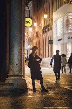 urban night scene that appears to be on a city street. A woman is standing in the foreground, checking something on her mobile phone. She is wearing a long coat and seems to be engaged with her device