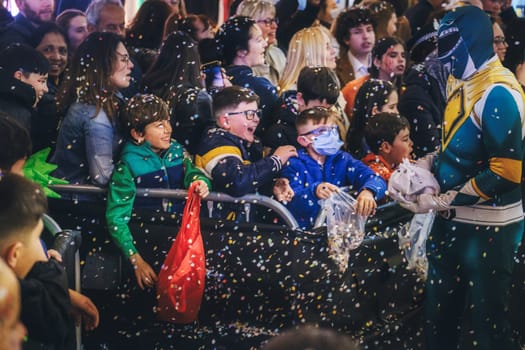 festive scene with a crowd of people, including children and adults, gathered likely at some sort of event or celebration. In the foreground, several children look excited as they are engaging with a person dressed in a colorful superhero costume. There is confetti in the air, adding to the celebratory atmosphere. The children appear to be delighted, with one of them holding a red bag