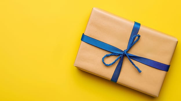 A brown gift box with a blue ribbon tied in a bow sits on a yellow background - Generative AI