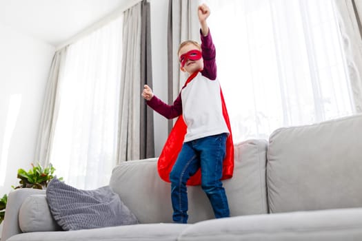 A boy dressed in a superhero costume with a red cape and mask standing on a grey couch. The child poses with arms raised triumphantly. The scene is set in a bright living room.
