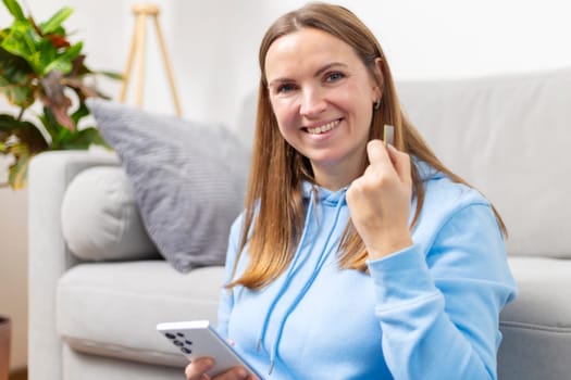 Smiling woman in blue hoodie sitting on a couch at home, holding a credit card and a smartphone. She appears to be making an online purchase or managing her finances.