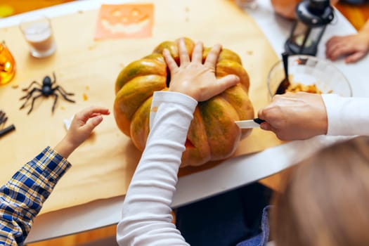 Family members are carving a pumpkin together at a table. One person holds the pumpkin steady while another uses a knife. Halloween decorations and tools are scattered on the table. Indoor setting.