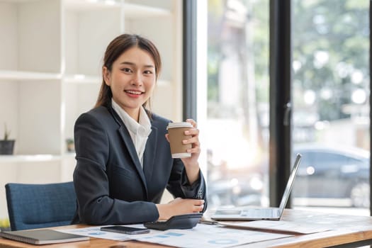 Photo of positive Asian businesswoman holds a cup of coffee imagines plan holiday in office..