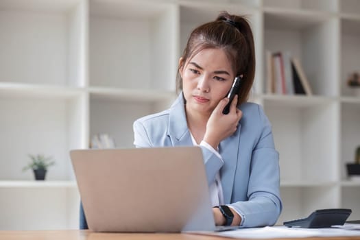 Attractive Asian woman using laptop in online meeting. Businesswoman using mobile phone talking with colleague sitting at table in modern office.