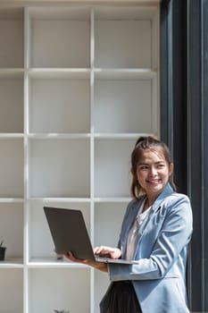 Portrait, Professional and confident Asian businesswoman or female executive manager in formal suit and use laptop. standing, leaning on table, holding laptop and using laptop computer..