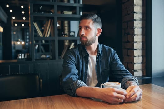 Confident man enjoying a cup of coffee while having work break lunch in modern restaurant, young intelligent man or entrepreneur relaxing in indoors cafe looking pensive