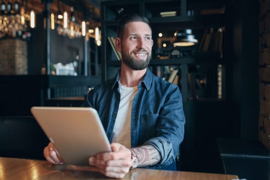 Modern hipster businessman drinking coffee in the city cafe during lunch time and working on tablet. Freelancer