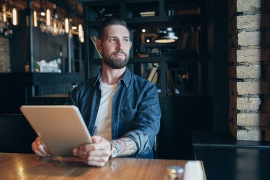 Modern hipster businessman drinking coffee in the city cafe during lunch time and working on tablet. Freelancer