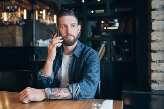 Talking with friends. Confident young man talking on the mobile phone and smiling while sitting in cafe