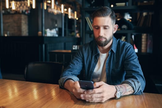 Young bearded businessman,dressed in a denim shirt, sitting at table in cafe and use smartphone. Man holding smartphone and looking at its screen. Man using gadget. Guy browsing internet on smartphone