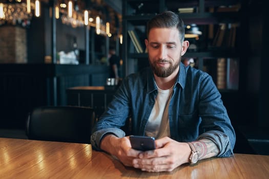 Young bearded businessman,dressed in a denim shirt, sitting at table in cafe and use smartphone. Man holding smartphone and looking at its screen. Man using gadget. Guy browsing internet on smartphone