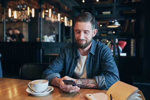 Young bearded businessman,dressed in a denim shirt, sitting at table in cafe and use smartphone. Man holding smartphone and looking at its screen. Man using gadget. Guy browsing internet on smartphone