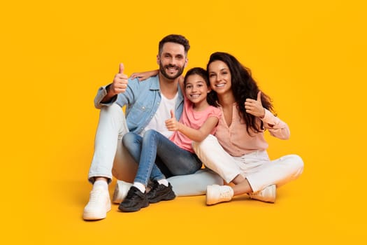 A happy family of three, a mother, father, and young daughter, sit on a yellow background, smiling and giving thumbs up. The family is dressed in casual clothes, they are all looking at the camera.