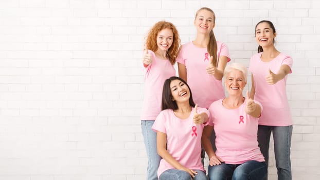 Diverse Female Volunteers In Pink Breast Cancer T-Shirts With Ribbons Gesturing Thumbs-Up Posing Over White Brick Wall. Copy Space