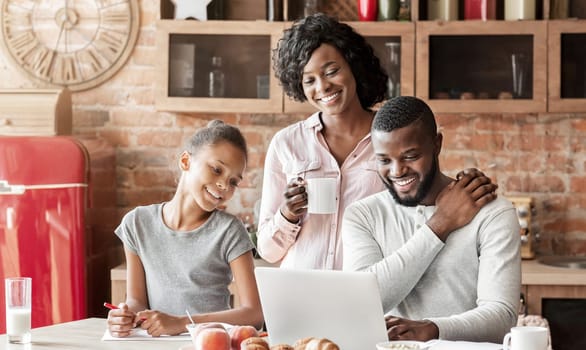 Lovely african family having breakfast, watching photos from last vacation on laptop, kitchen interior, copy space
