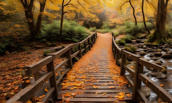 A wooden bridge with railings extends through an autumn forest, leading to a pathway covered in fallen leaves. A small stream runs along the edge of the bridge, with stones scattered along its banks.