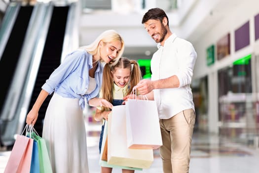 A young girl is standing between her parents, all three looking down at a pile of shopping bags. The girl is smiling, and the parents are smiling as they look at the contents of the bags.
