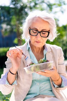 portrait of a middle age woman eating a takeaway salad sitting outdoors in a city park, concept of healthy food and active lifestyle in adulthood, copyspace for text