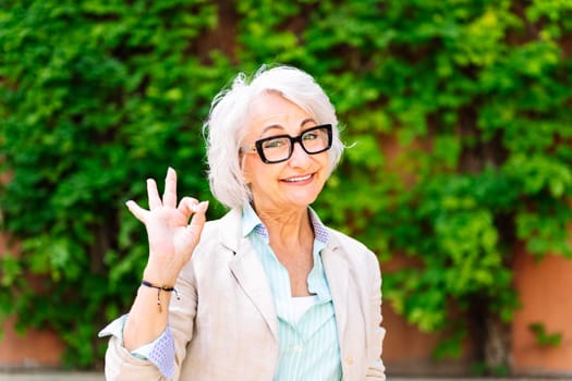 portrait of a middle age woman smiling happy and doing ok sign with hand while looking at camera with blurred green plants background, concept of mature people lifestyle