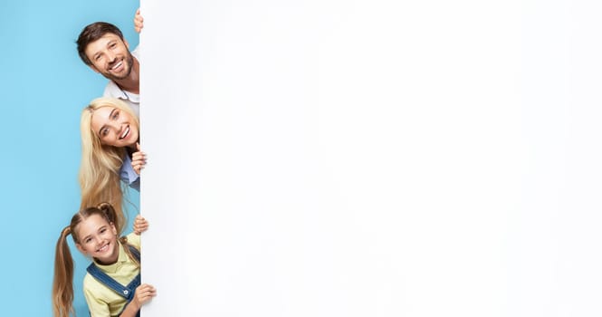 A family of three smiles brightly as they peek from behind a large white wall. The father stands at the top, followed by the mother, and their young daughter.
