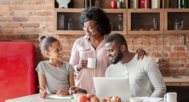 Quality time together. Happy african american family spending morning at kitchen, having breakfast, girl painting, daddy using laptop, copy space