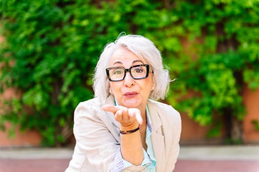 portrait of a middle age woman blowing a kiss with her hand while looking at camera with blurred green plants background, concept of mature people lifestyle