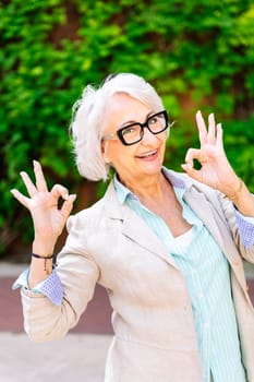 portrait of a middle age woman smiling happy and doing ok sign with hands while looking at camera with blurred green plants background, concept of mature people lifestyle
