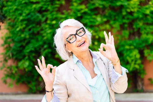portrait of a middle age woman smiling happy and doing ok sign with hands while looking at camera with blurred green plants background, concept of mature people lifestyle