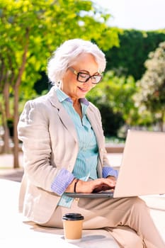middle age woman smiling happy using laptop sitting outdoors in a city park, concept of technology and modern mature people lifestyle