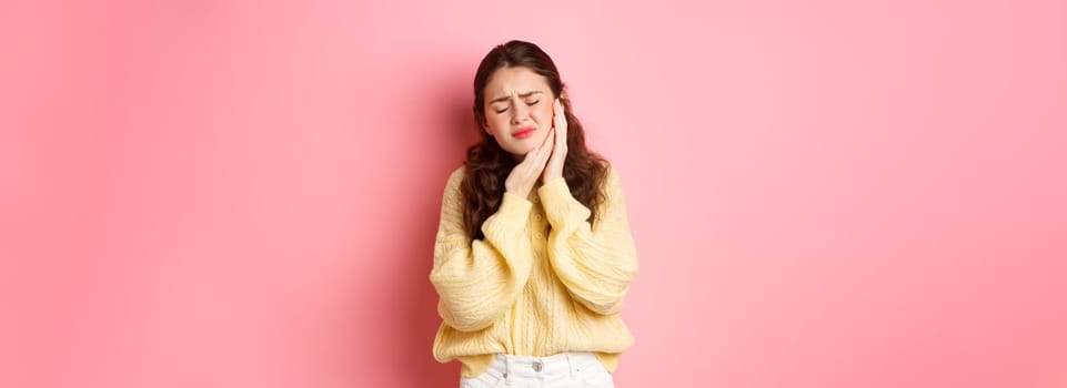 Young woman with terrible toothache, touching swallen cheek and grimacing from painful feeling, having tooth decay, need appointment to dentist, standing against pink background.