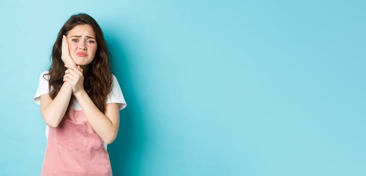 Image of frowning upset girl touching swallen cheek, having toothache, complaining on tooth decay, need to see dentist, standing against blue background.