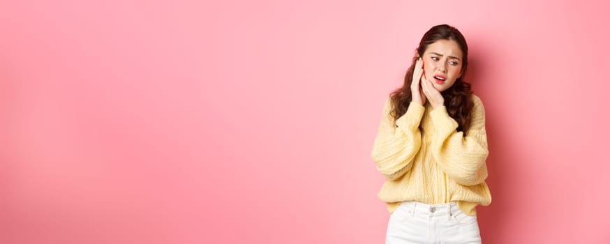 Young woman having toothache, touching swallen cheek and grimacing from pain, need to see dentist, stomatology appointment, standing against pink background.