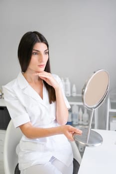Confident dermatologist holding mirror at desk. Happy female cosmetologist working in modern medical office. Woman in white uniform sitting near in clinic.