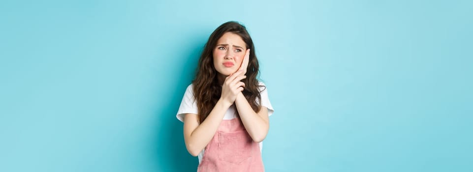 Portrait of frowning girl touching cheek, having toothache, looking at upper left corner stomatology clinic banner, suffering tooth decay, standing against blue background.