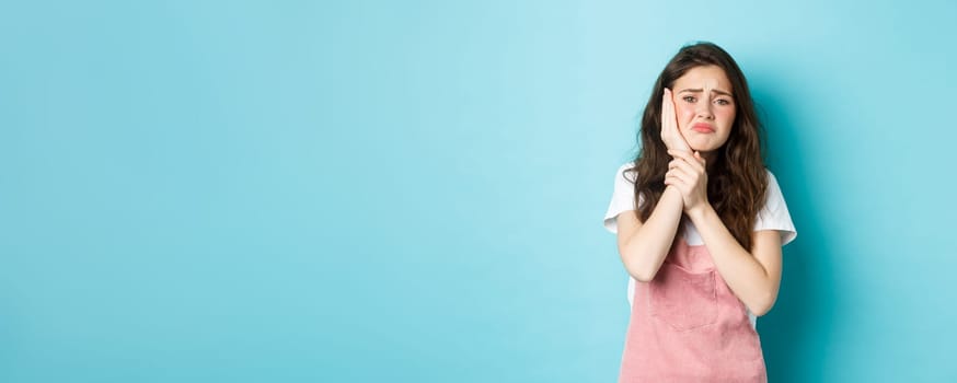Image of frowning upset girl touching swallen cheek, having toothache, complaining on tooth decay, need to see dentist, standing against blue background.