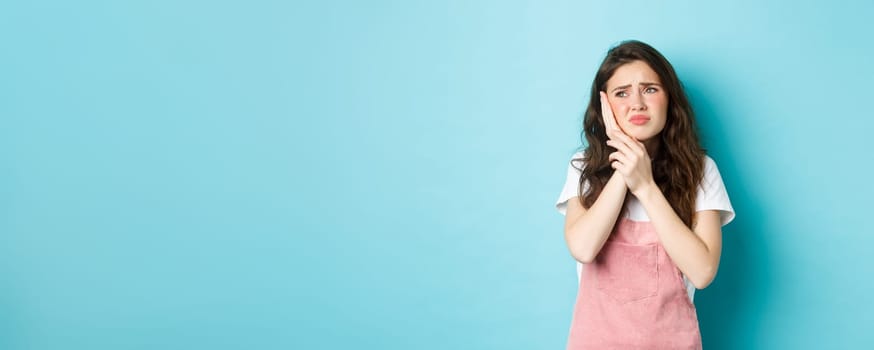 Portrait of frowning girl touching cheek, having toothache, looking at upper left corner stomatology clinic banner, suffering tooth decay, standing against blue background.