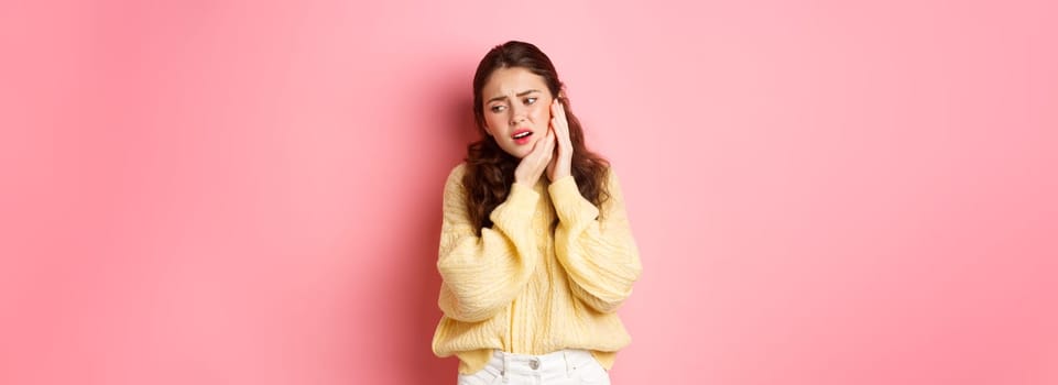 Young woman having toothache, touching swallen cheek and grimacing from pain, need to see dentist, stomatology appointment, standing against pink background.