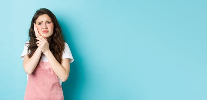 Portrait of frowning girl touching cheek, having toothache, looking at upper left corner stomatology clinic banner, suffering tooth decay, standing against blue background.