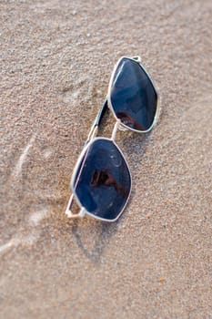 Sunglasses lie on the wet sand on the sea beach, with the reflection of a beautiful girl in the glass of the glasses, on a sunny summer day,