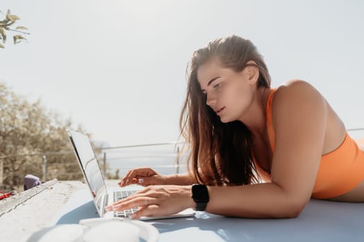 A woman in an orange sports bra lies on her stomach while working on a laptop. She has long brown hair and is looking at the screen.
