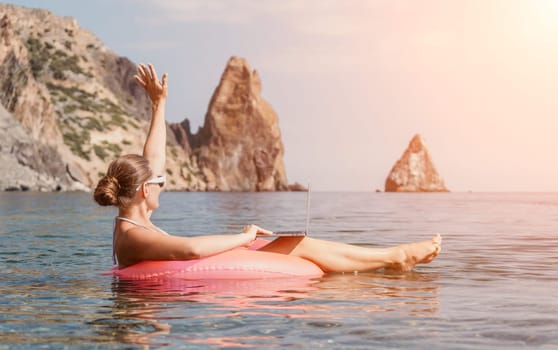 Woman freelancer works on laptop swimming in sea on pink inflatable ring. Pretty lady typing on computer while floating in the sea on inflatable donut at sunset. Freelance, remote work on vacation