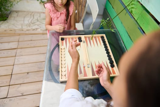 A mother and her daughter engage in a fun game of backgammon at an outdoor table. The family bonding moment is highlighted in a natural, sunny setting.