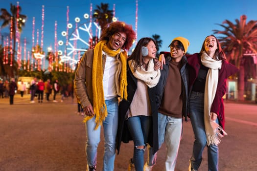 Group of happy multiracial friends walking around city laughing and talking together on winter night near Christmas market. College student and lifestyle concept.