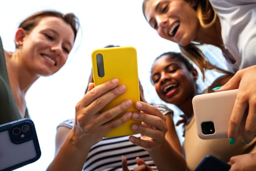 Low angle view of happy multiracial group of young women friends looking at mobile phone. Youth lifestyle and social media concept.
